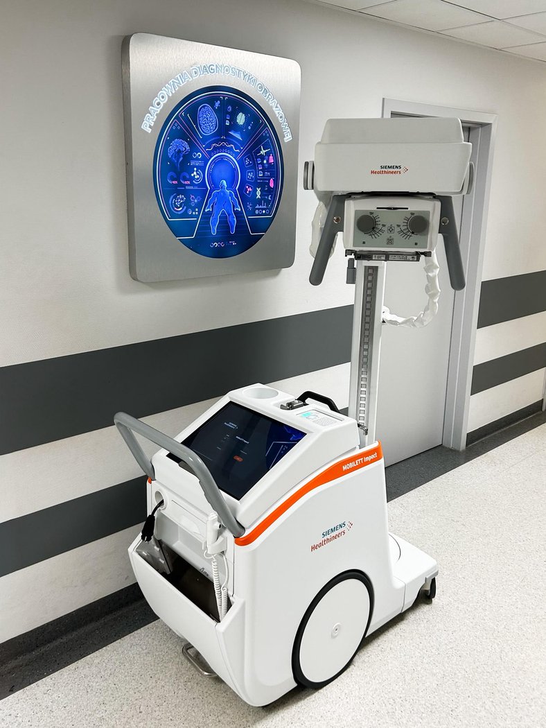 Mobilny aparat rentgenowski Siemens Healthineers stojący na korytarzu przed wejściem do pracowni diagnostyki obrazowej. W tle na ścianie znajduje się podświetlany znak z napisem „Pracownia Diagnostyki Obrazowej” oraz grafiką przedstawiającą sylwetkę człowieka i motywy medyczne